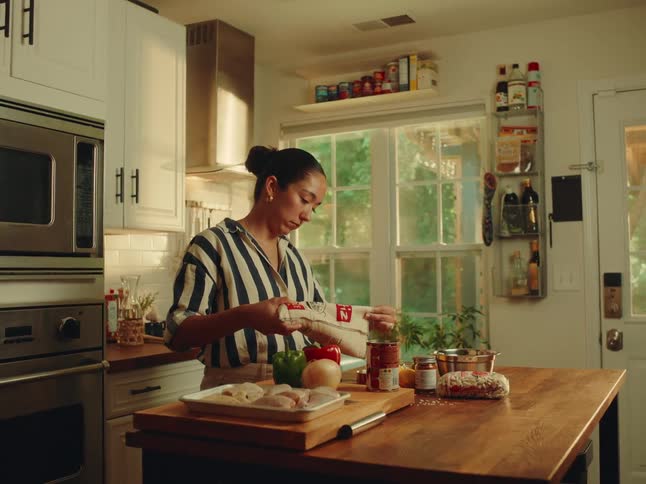 A woman with her hair in a bun wearing a striped shirt is focused on opening a package while standing at a wooden island counter laden with various food ingredients in a wellequipped kitchen with white cabinets and a large window