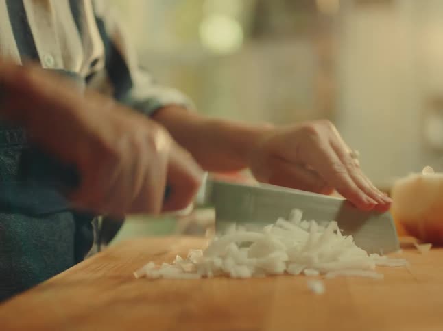 A closeup shot shows a persons hands skillfully dicing an onion on a wooden cutting board with finely chopped pieces already accumulated the person is wearing a striped shirt and an apron suggesting they are in the midst of food preparation