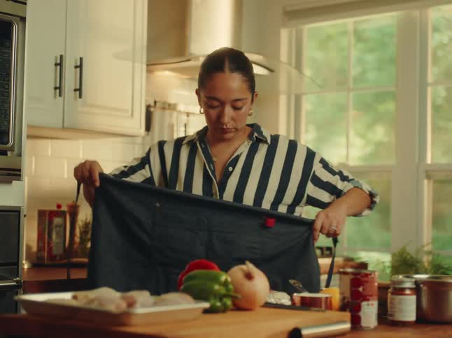 A woman with her hair pulled back wearing a navy and white striped shirtputting an apron and there ingredients on a wooden counter in a brightly lit kitchen with white cabinets and a large window