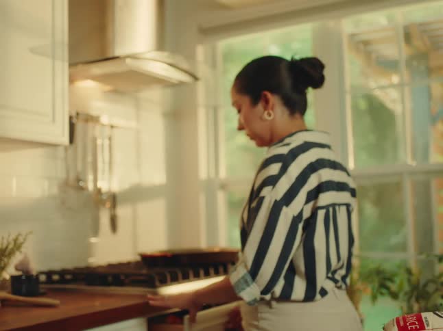 The camera focuses on fresh ingredients on a wooden countertop including green and red bell peppers an onion and raw chicken pieces in a tray in the background a person in a striped shirt is seen moving around a welllit kitchen with a large window