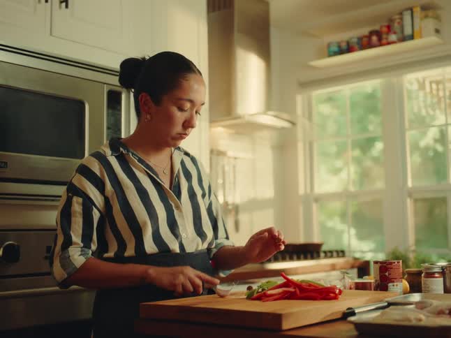 A young adult woman with her hair in a bun wearing a striped shirt and an apron intently chops vegetables on a wooden cutting board in a brightly lit kitchen with a window looking out at trees