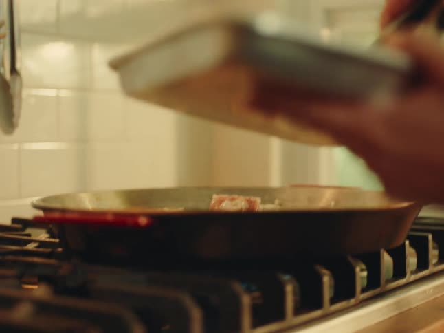A person uses tongs to sear meat in a skillet on a stovetop