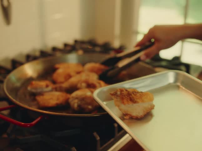 A hand using tongs to transfer browned chicken pieces from a large pan on a stove to a metal baking sheet