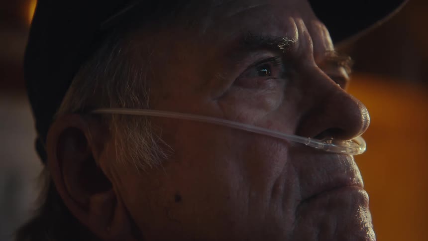 An elderly man wearing a nasal bridle and a hat is sitting indoors looking sad