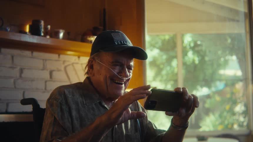 Elderly man with a small oxygen tube in his nose with a happy expression sitting in a wheelchair and interacting with a smartphone
