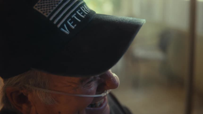 The video shows a closeup profile of an elderly man wearing a veteran baseball cap and an oxygen tube he appears to be smiling and looking up with a soft blurry background suggesting an indoor setting