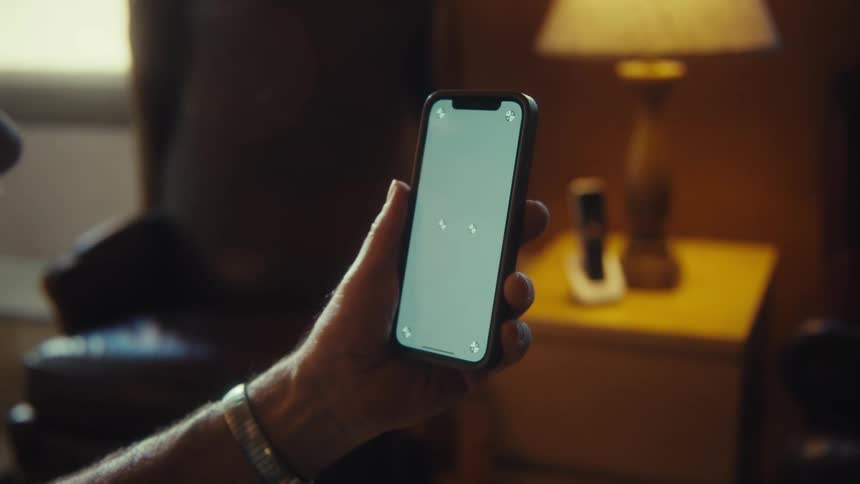 A closeup shot of a persons hand holding a smartphone with a blank lightcolored screen and tracking markers on all four corners the camera slowly zooms out revealing the person is sitting in a room with a lamp and a chair in the background