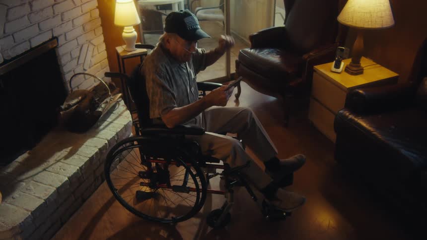 An elderly man wearing a baseball cap and glasses is seated in a wheelchair in a dimly lit room illuminated by the warm glow of lamps he holds a phone and shows signs of excitement gesturing with his free hand suggesting hes reacting to content on
