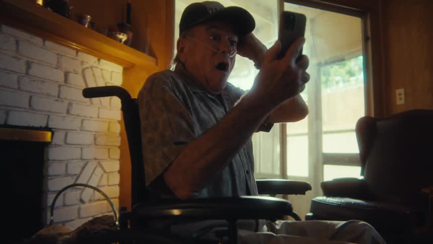 An elderly man wearing a baseball cap and with a nasal cannula visible is seated in a wheelchair in a sunlit room he holds a smartphone first appearing focused on it then raising his arm and expressing excited surprise possibly in response to someth