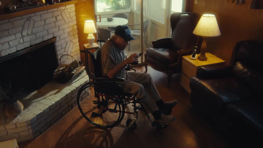 The video shows a man in a wheelchair holding a remote control he is sitting in a living room with a fireplace and two lamps