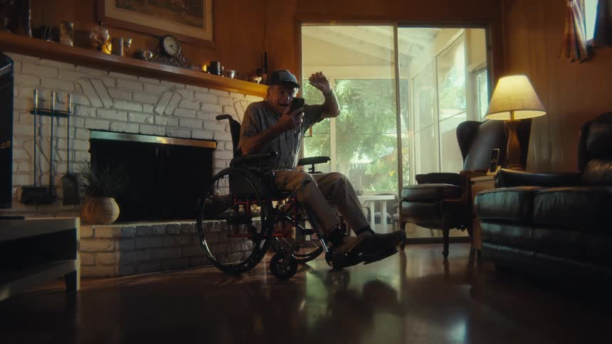 An elderly man wearing a hat sits in a wheelchair overly excited by the content hes watching on his phone in his living room at midday