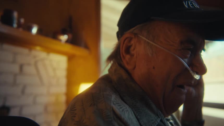An elderly man with a nasal bridle is overly excited while watching a program on his phone seated in a wheelchair at midday
