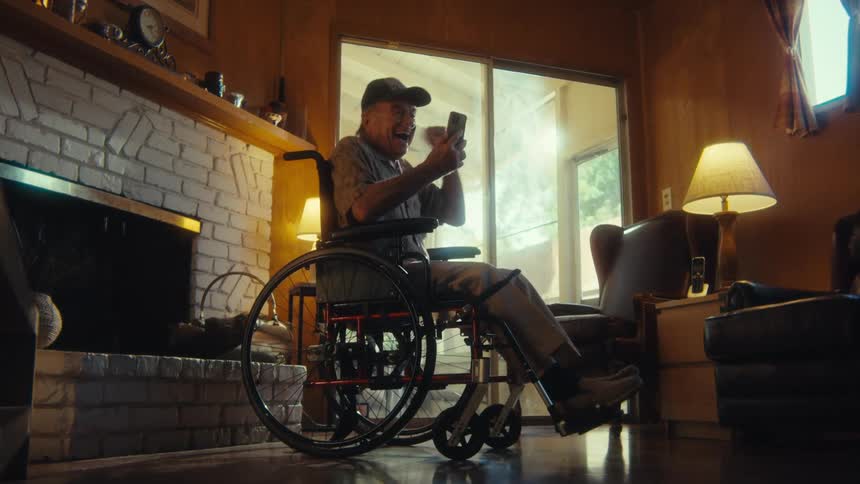 An elderly man wearing a hat and a nasal bridle sits in a wheelchair overly excited by the content he is watching on his phone