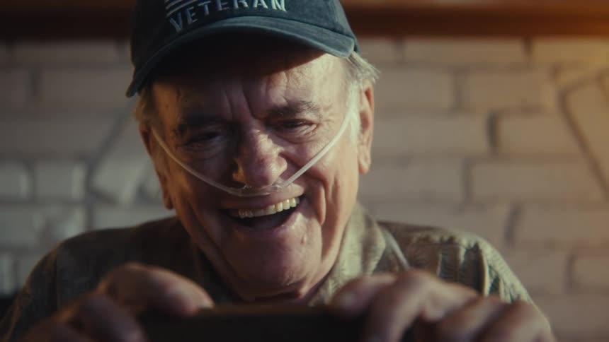 The video shows a closeup of an elderly man with an oxygen tube in his nose wearing a veteran cap and smiling widely while looking at a smartphone the background slowly widens to reveal a living room with a fireplace and mantle