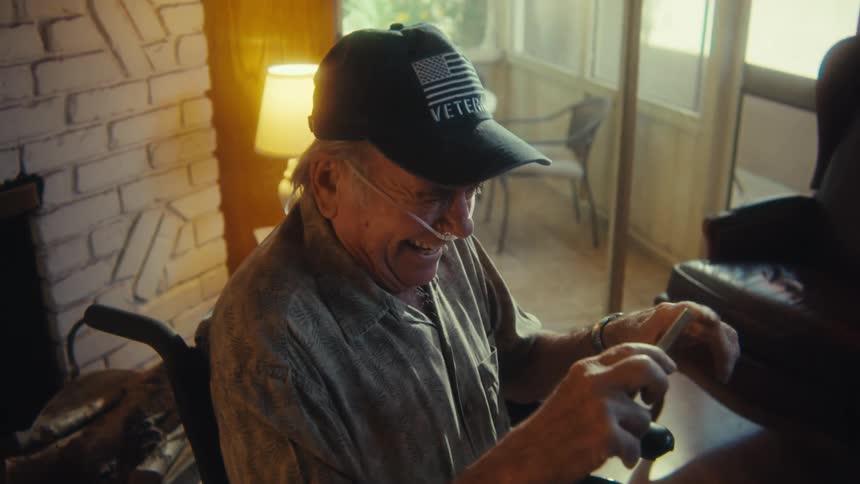 An elderly man wearing a veterans cap is seated in a wheelchair actively engaged with a small device in his hands possibly a phone or remote control he looks up and raises his arm in a gesture of celebration or excitement then returns his focus to t