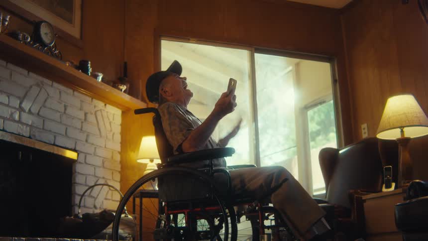 An elderly man wearing a baseball hat sits in a wheelchair indoors with a nasal bridle on his nose overly excited by the content hes watching on his phone held in his right hand seated in his living room beside the fireplace