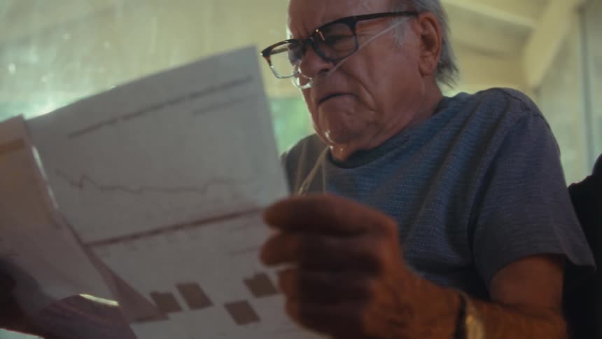 The video shows a man with glasses and an oxygen tube looking at a document with graphs he appears to be examining the paper with a focused expression