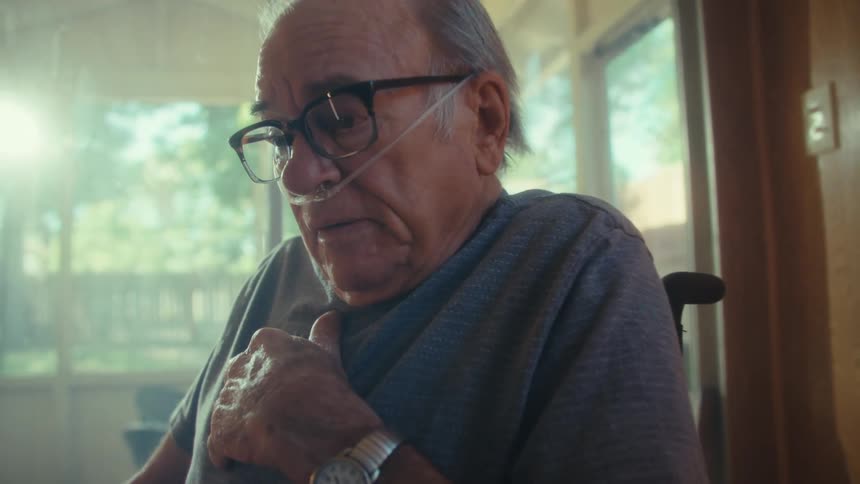 An elderly man sitting in a wheelchair beside a glass window wearing reading glasses and a nasal bridle reading a document