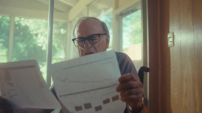 An elderly man wearing reading glasses sits in a wheelchair frowning as he reads a document with a nasal bridle attached to his nose