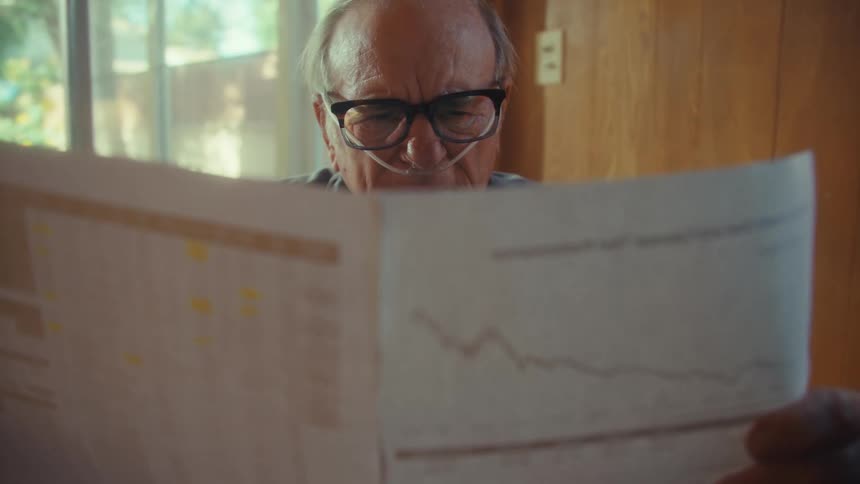 An elderly man wearing reading glasses sits beside a glass window reading a document that leaves him looking sad with a nasal bridle attached to his nose
