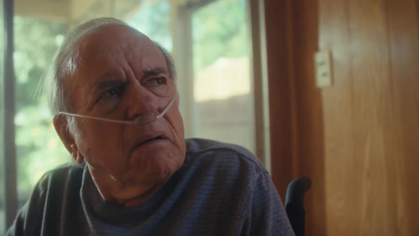 An older adult man with light colored hair wearing glasses and a nasal cannula sits holding a piece of paper looking forward with a serious expression and then looks up with a worried gaze inside a room with a large bright window