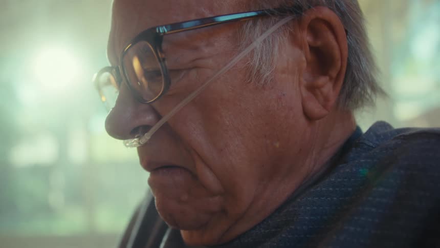 An elderly gentleman with light colored hair and a nasal cannula looks down with a solemn expression then puts on his glasses and looks forward in a brightly lit room