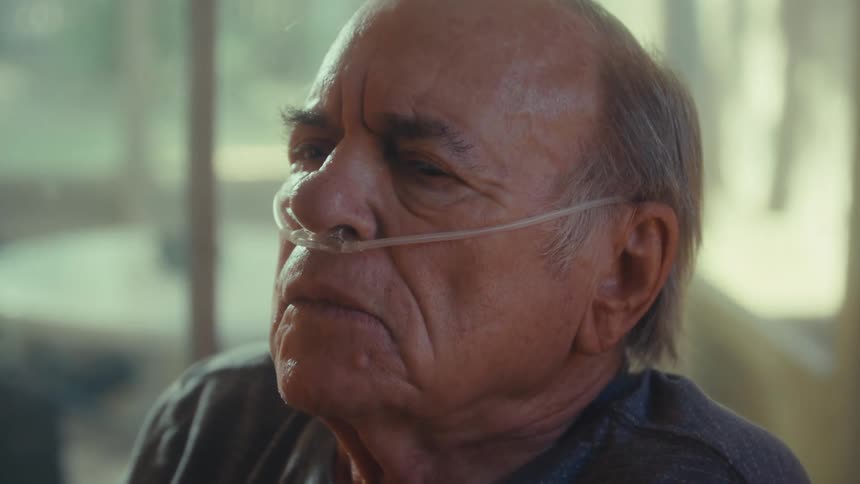 An elderly man sitting in a wheelchair with a nasal bridle on his nose beside a window taking a deep breath with his eyes closed then slowly opening them afterward