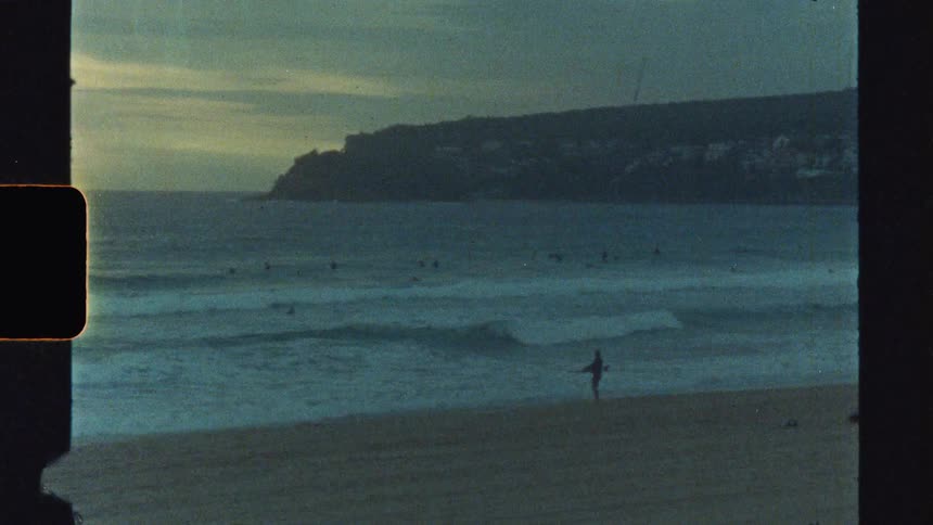 A person stands in the shallow ocean water with a surfboard at a sandy beach with waves breaking towards the shore and a dark tree covered cliff in the background under a cloudy sky