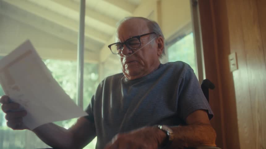 An elderly man sits in a wheelchair looking sad after reading a document