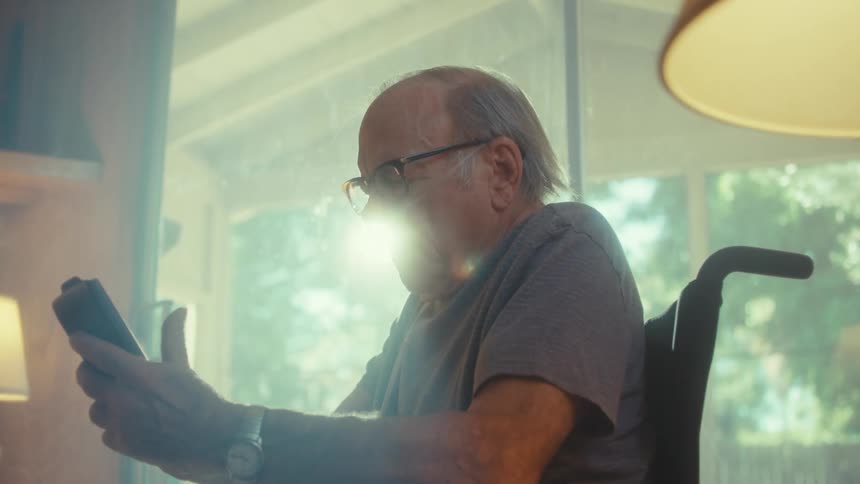 An elderly man wearing reading glasses sits in a wheelchair beside a window indoors scrolling through his phone with a nasal bridle attached to his nose
