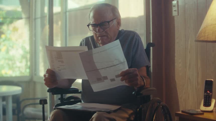 An elderly man sitting in a wheelchair indoors beside a glass window at midday reading a document he suddenly looks surprised and takes off his reading glasses with a nasal bridle on his nose