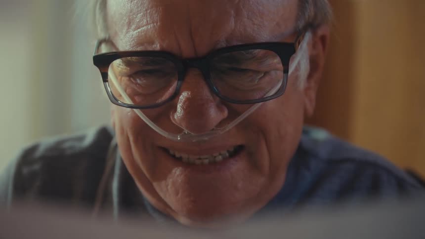 An elderly man wearing reading glasses sits while going through some documents in his hands he suddenly becomes sad with a nasal bridle on his nose