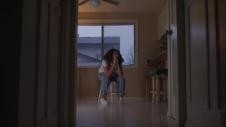 A teenage girl with long curly hair sits on a stool indoors resting both hands on her chin deep in thought