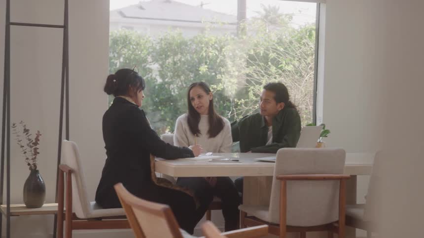 Three people are sitting at a white table and the woman in the middle is talking while the man next to her is nodding his head