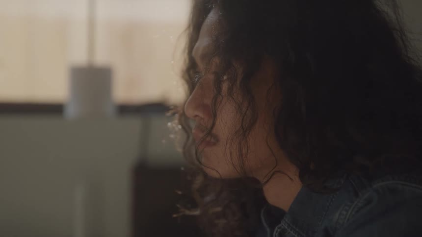 A young man with long curly hair sits indoors appearing calm and relaxed