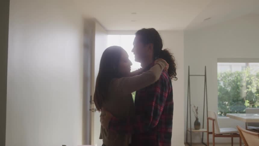 A young man and woman hug each other in a living room at midday with the door leading outside open