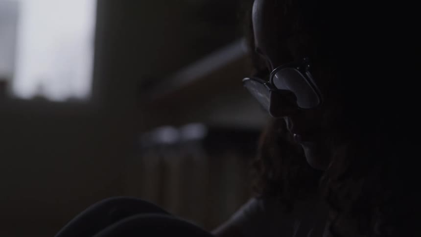 The face of a young woman wearing glasses who is sitting on the floor of a partially dark room