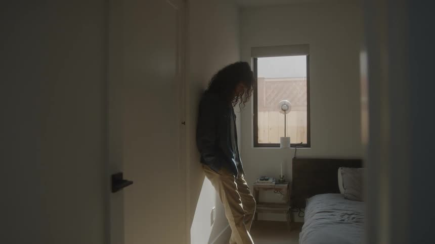 Curlyhaired man leaning against the wall of a room in the house looking dejected