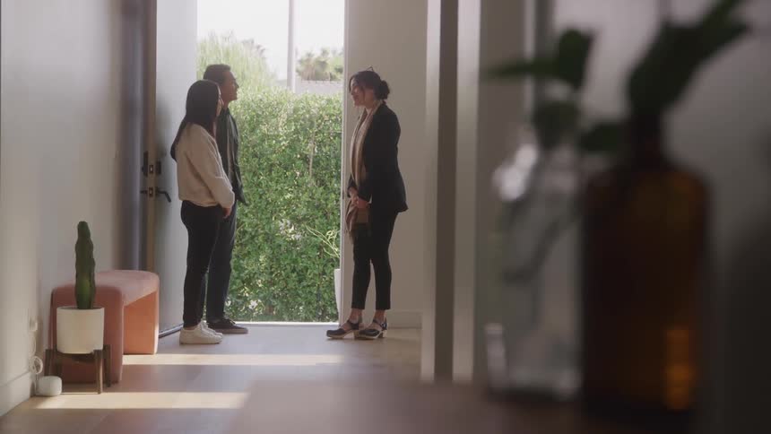 A woman in black clothes and a yellow scarf is greeting a couple who are entering through the door of a house with white walls