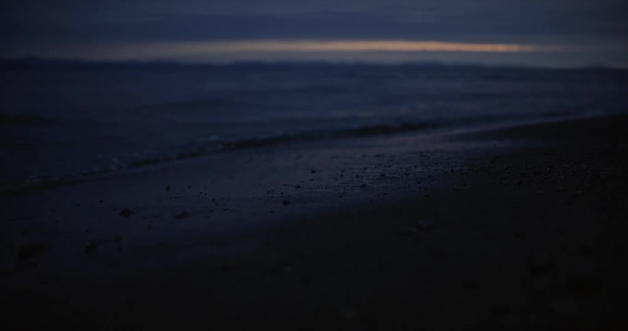 The video shows a dark dusky scene of waves gently lapping onto a sandy shore a faint band of orange light is visible on the horizon suggesting either sunrise or sunset