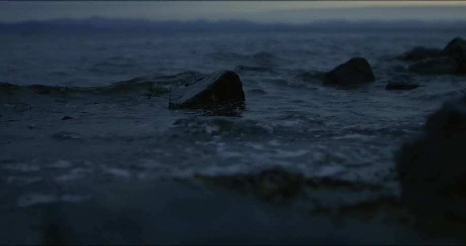A moody closeup shot of dark choppy waves crashing against rocks under a dim sky