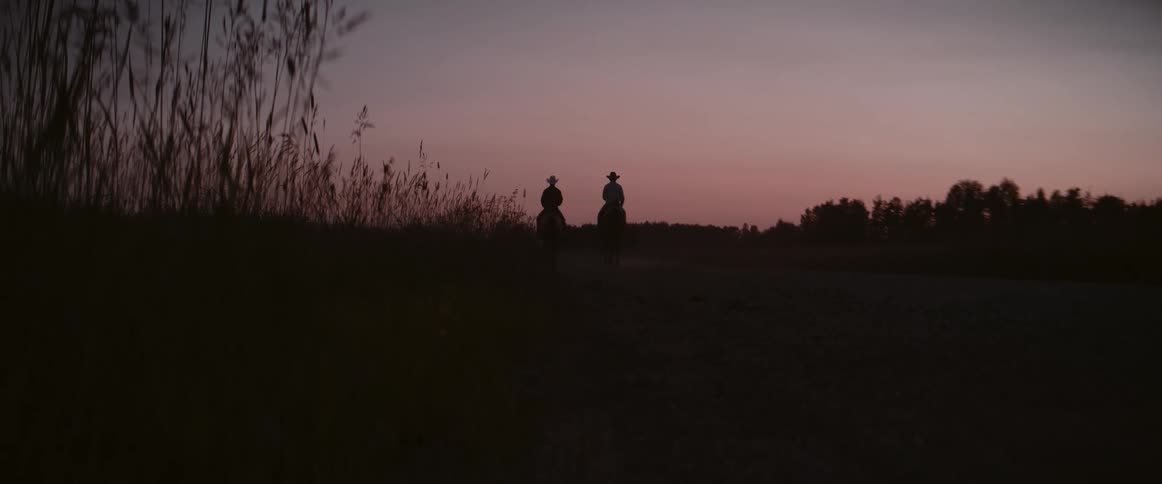 Two riders on horseback travel side by side along a quiet path at dusk silhouetted against the fading sky