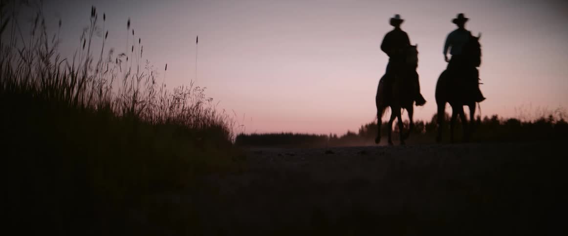 Two individuals on horseback are silhouetted against a colorful sunset sky riding on a dirt path with tall grasses in the foreground