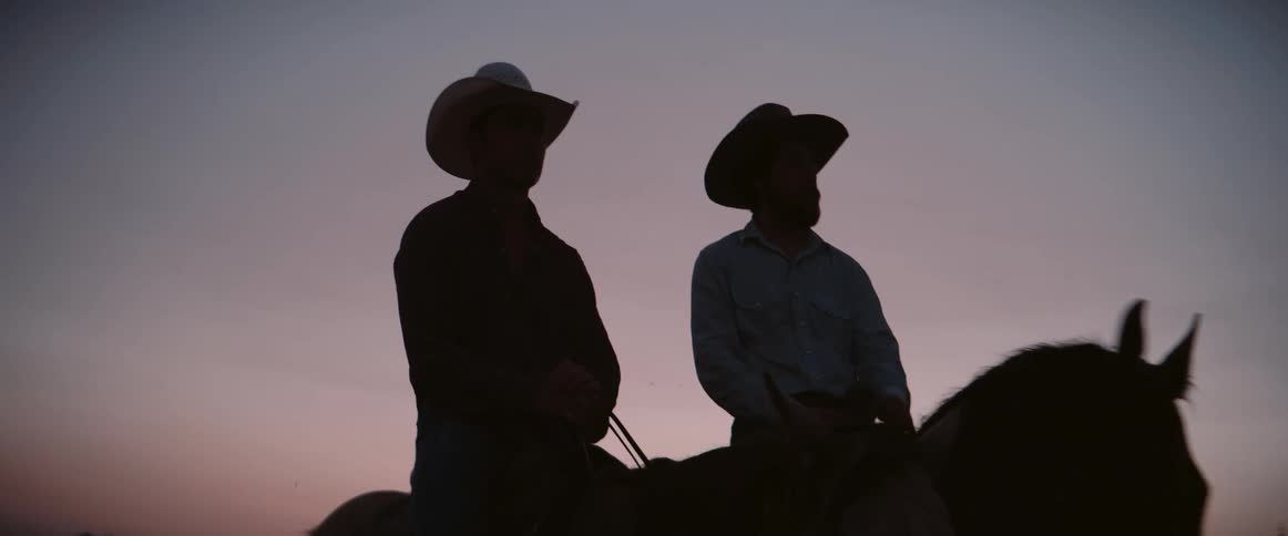 Two cowboys ride horses together during sunset