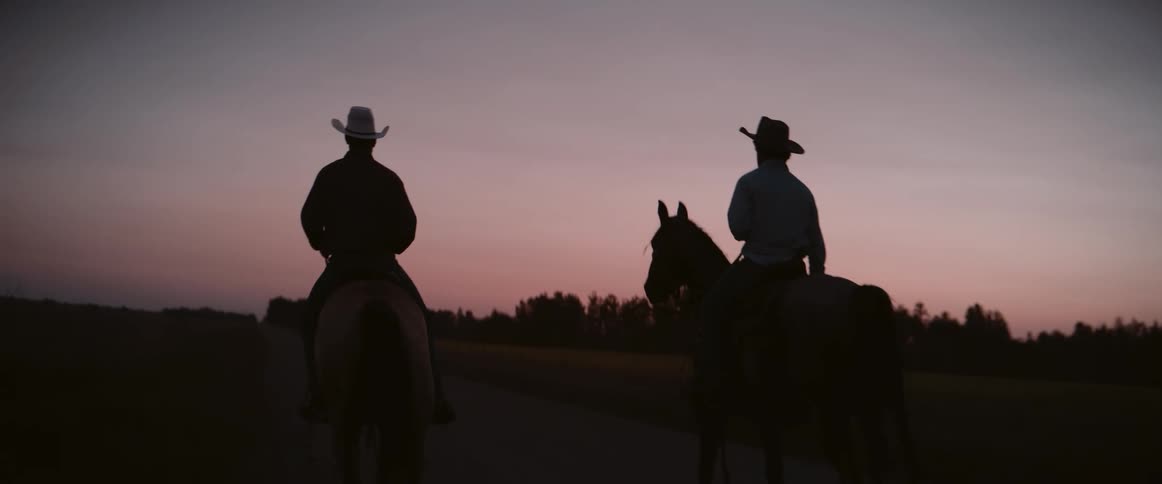 Two cowboys ride side by side at sunset their silhouettes sharply outlined against the fading light