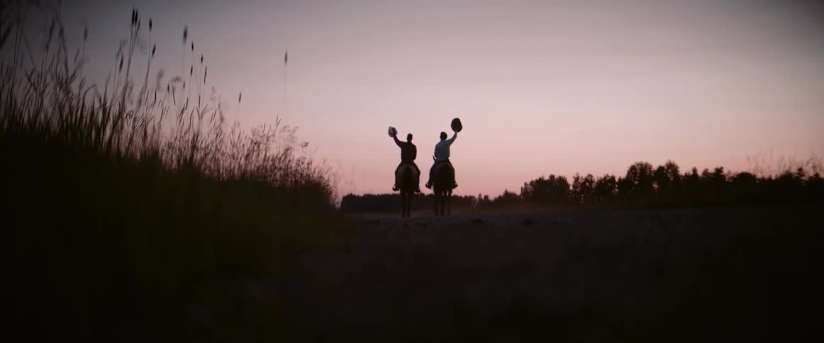 Two male figures on horseback wearing cowboy hats ride away from the camera on a dirt path during sunset silhouetted against the colorful sky