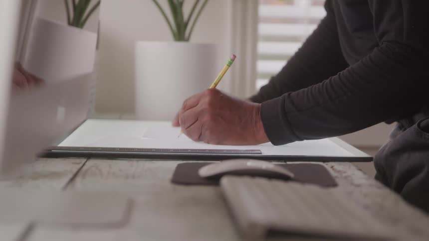 A person is shown from the chest down wearing a black longsleeved shirt and drawing on a light tablet with a pencil the setting appears to be an office or creative workspace