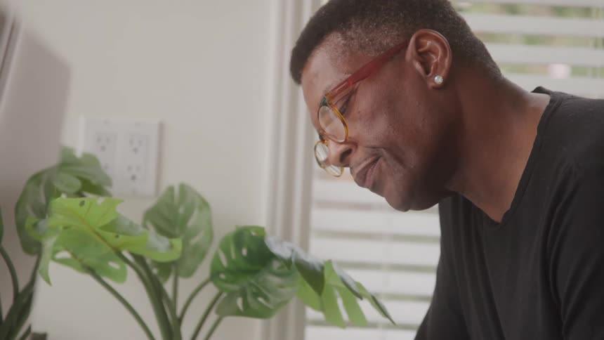 A closeup side view of a man with glasses intently looking at a green houseplant indoors