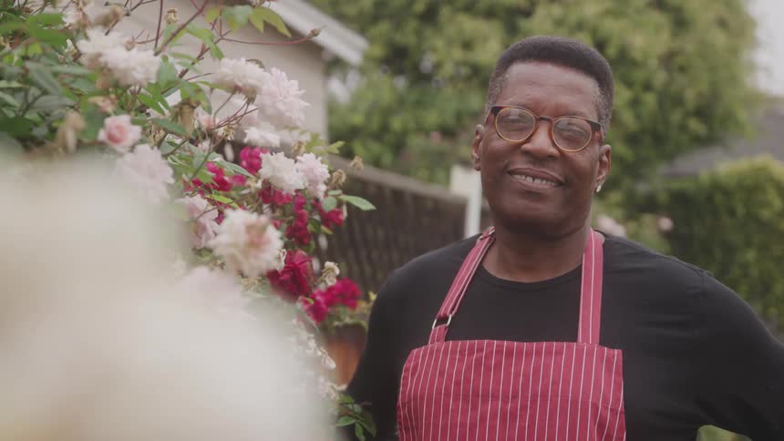 Man in an apron stands next to a flower bush in a garden