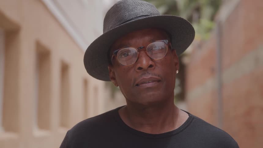 A middleaged man is outdoors at midday wearing a hat clear glasses and a black tshirt